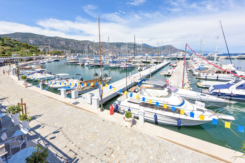 BEAULIEU-SUR-MER, FRANCE - JUNE 28, 2017: Daylight sunny view to parked yachts in port . People and childrens walking on sidewalk near boats. Mountains and bright blue sky with clouds on background.